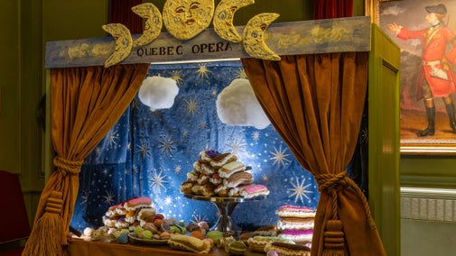 A large model of an opera stage in the Bicentennial Room at Quebec House. Crocheted macarons and eclairs fill the stage. Stars and clouds form the backdrop. Smiling moon cycles at the top of the stage. A portrait of General James Wolfe hangs on a wall in the background.